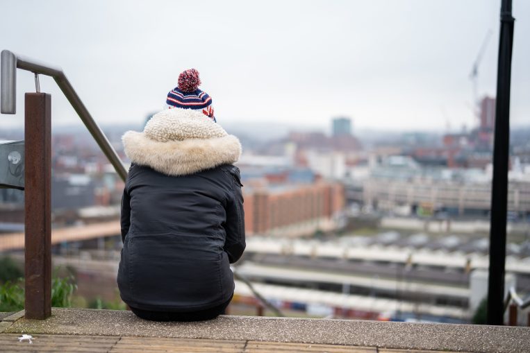 Young person with a winter coat on sat on stairs looking over a city