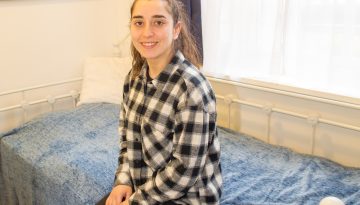 Young woman sat on a single bed with her hands on her lap smiling at the camera