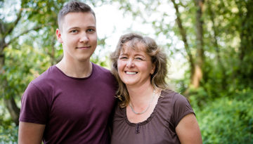 Older woman and boy smiling looking directly to the camera. In the background there are blurred trees