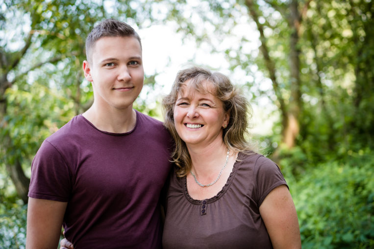 Older woman and boy smiling looking directly to the camera. In the background there are blurred trees