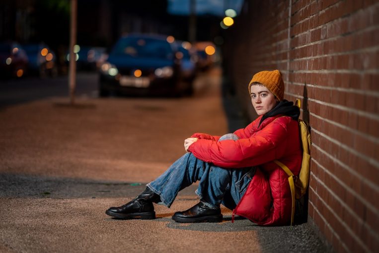Young homeless woman sitting on the street resting against a brick wall. She is wearing a red coat and yellow beanie.