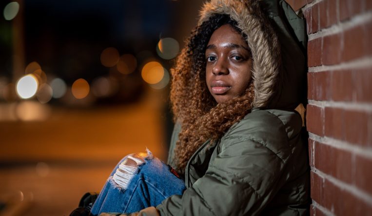 Young homeless woman sitting on the street with her hood up. She is looking directly at the camera.