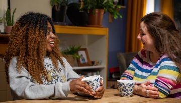 Young women and older woman smiling at each other drinking tea in a kitchen.