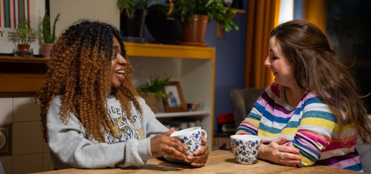 Young women and older woman smiling at each other drinking tea in a kitchen.