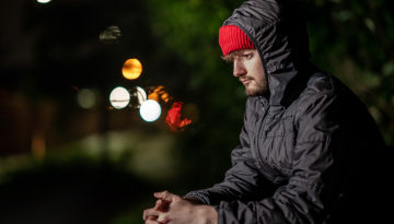 Young man sitting with hands together looking at the ground. He is wearing a padded coat and red hat.