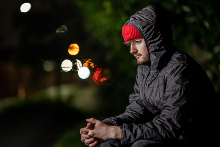 Young man sitting with hands together looking at the ground. He is wearing a padded coat and red hat.