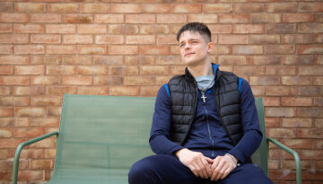 Young man sat on a garden bench in front of brick wall. Looking away from the camera