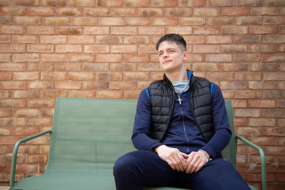 Young man sat on a garden bench in front of brick wall. Looking away from the camera